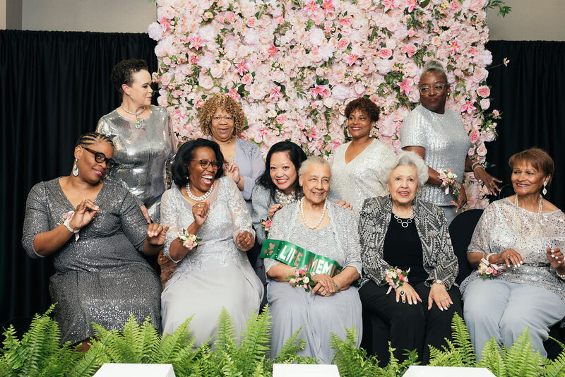Phi Alpha Omega Charter Members laughing and wearing silver gowns in front of a flower wall