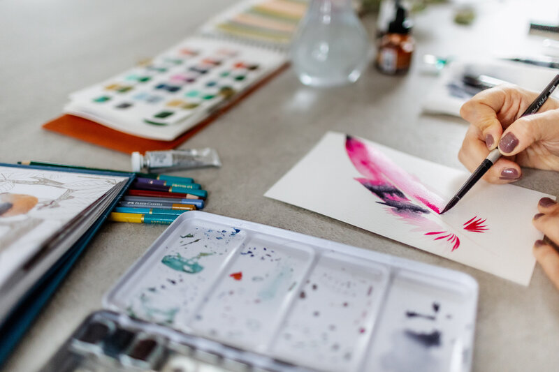 A watercolor artist’s hand painting a feather, surrounded by her painting tools.