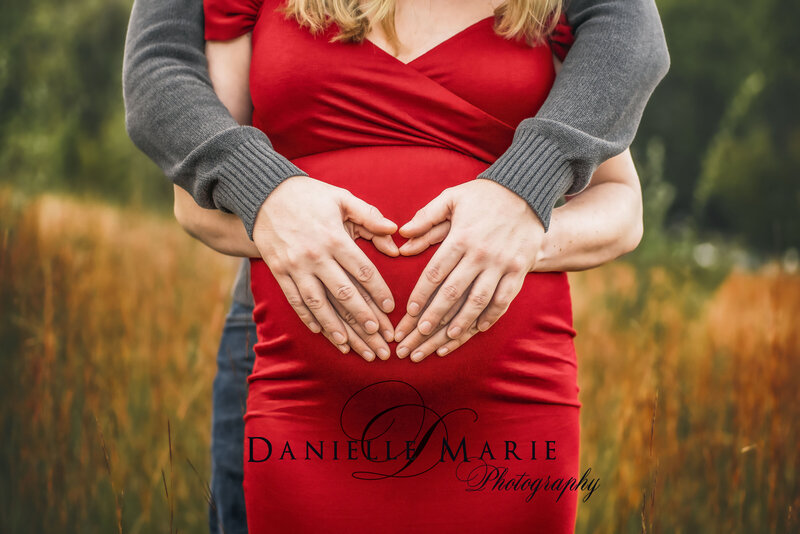 couple posing in open field while wearing a red dress and holding a heart over her baby bump in New Jersey 