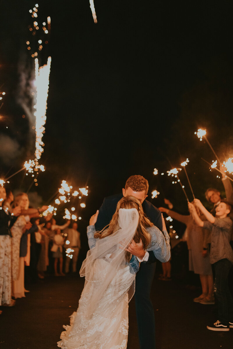 A newlywed couple share a kiss surrounded by friends holding sparklers.