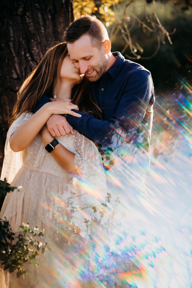 A couple hugs and kisses close together in front of a tree with a prism effect in the bottom corner of the image