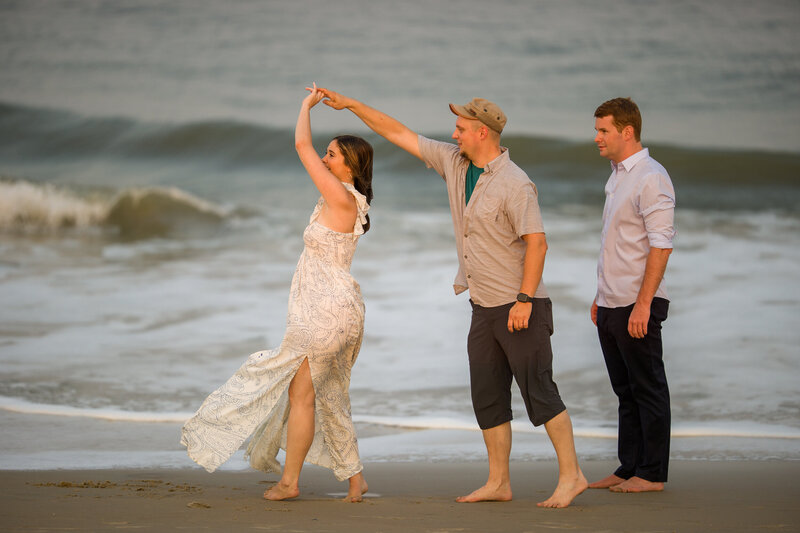 Travis Stitt demonstrating a playful spin pose on the beach for an engaged couple during their engagement session, capturing dynamic movement and creating fun, memorable moments by the ocean.