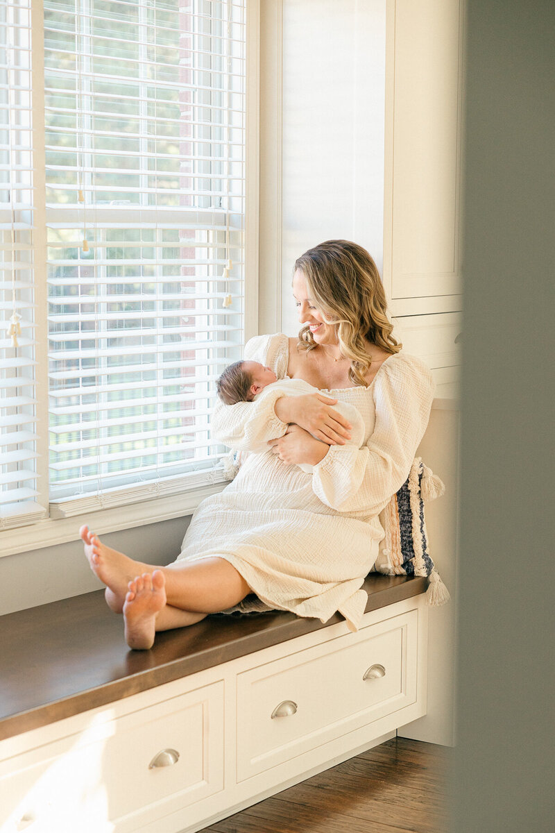 mother relaxes on a bench while holding her newborn baby dressed in cream and smiling down at him, Indianapolis newborn photographer