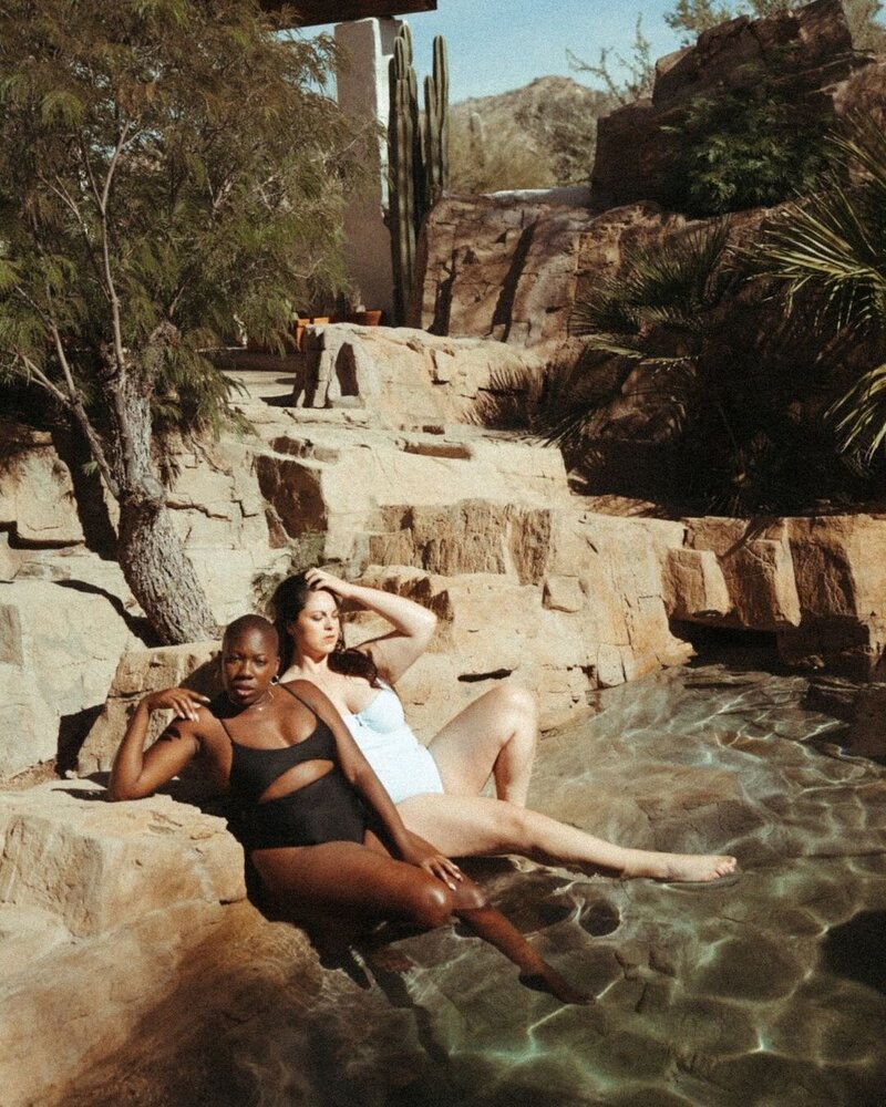 two women in swimsuits sitting in water surrounded by rocks