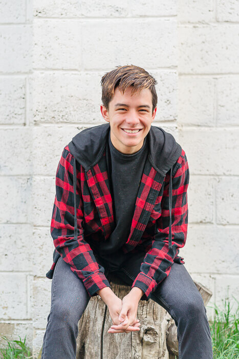 A high school senior boy wearing a red plaid jacket sits on a tree stump in front of a white brick wall in Lehi in the spring. Captured by Salt Lake City senior photographer Melissa Woodruff Photography