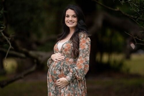 A-smiling-pregnant-woman-stands-outdoors-cradling-her-belly-and-wearing-a-floral-dress