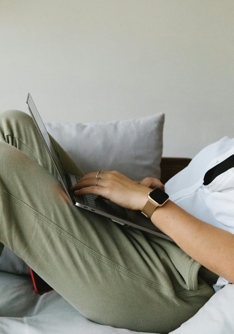 hands typing on a laptop seated on a sofa featuring a watch white button down green pants