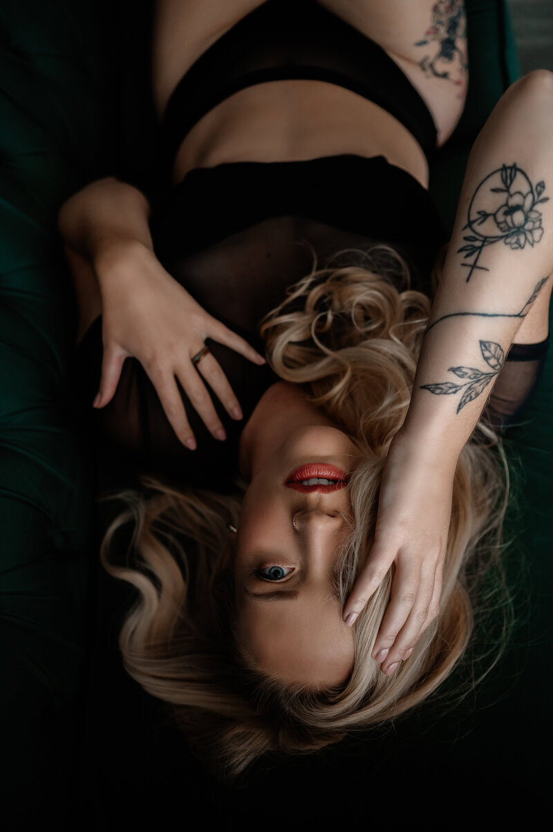 Up close boudoir shot of blonde woman wearing black lingerie laying elegantly on green couch with hands resting on her face delicately. Lips parting slightly and eyes staring into the camera creates a striking mood.
