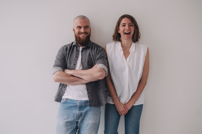 Marisa and Tanner of Msav Creative Co lean against a wall together, Marisa is laughing while Tanner is smiling with his arms crossed