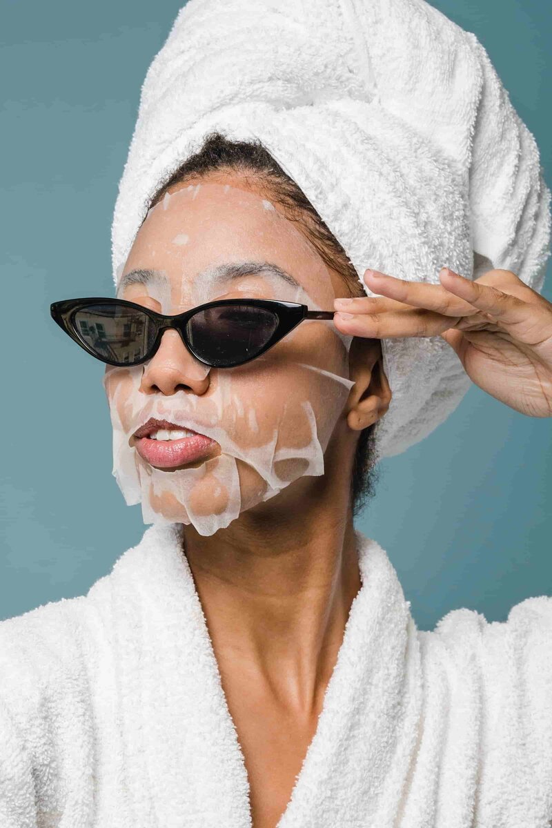 Woman in hydrating sheet mask while wearing a robe and sunglasses
