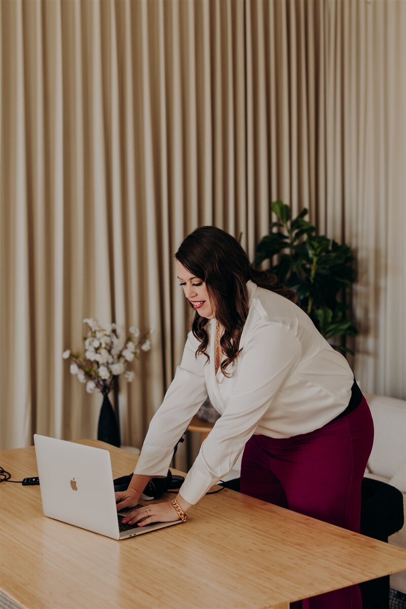 woman leaning over desk in white shirt and red pants typing on macbook