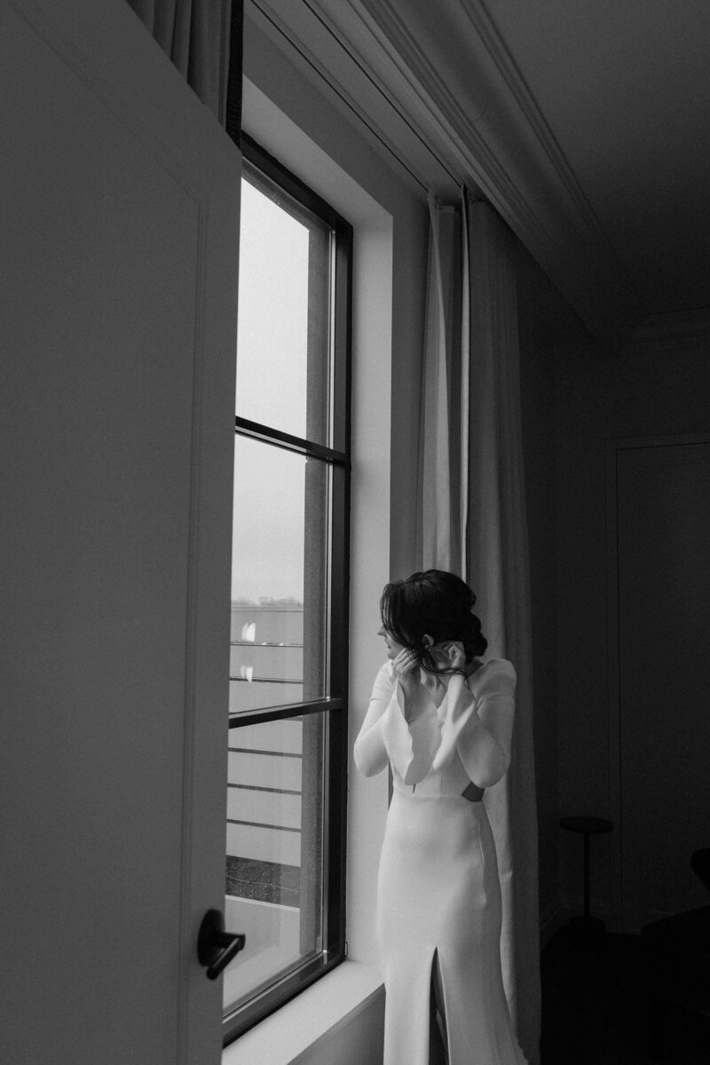 A person in a wedding dress putting on earrings and looking out a window 