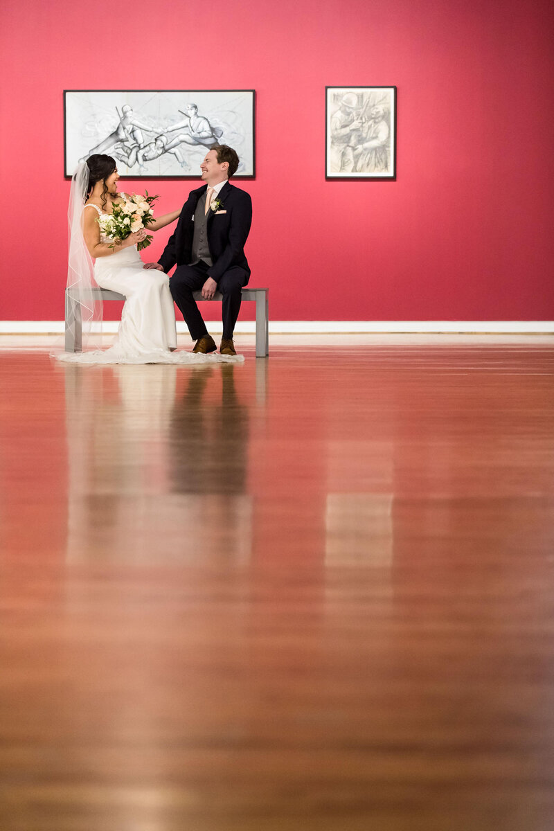 4-radiant-love-events-bride-groom-sitting-on-bench-laughing-in-distance-shot-upward-from-wooden-floor-art-on-red-wall-behind-them-romantic-elegant-timeless