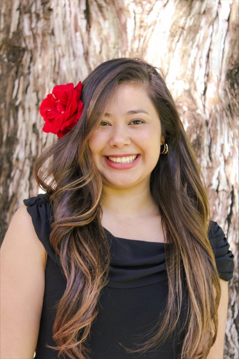 Doris, a Latina woman with long brown hair decorated with a red rose, smiles happily at the camera. She is wearing a black shirt and gold earrings and is framed by a large tree in the background.