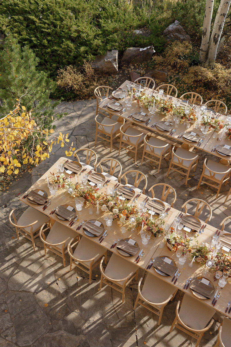 Image shows a  view from above the wedding reception dinner tables. Pale pine wood tables are set with dark brown chargers wrapped in mocha velvet napkins. Netural flowers dot the tablescape.