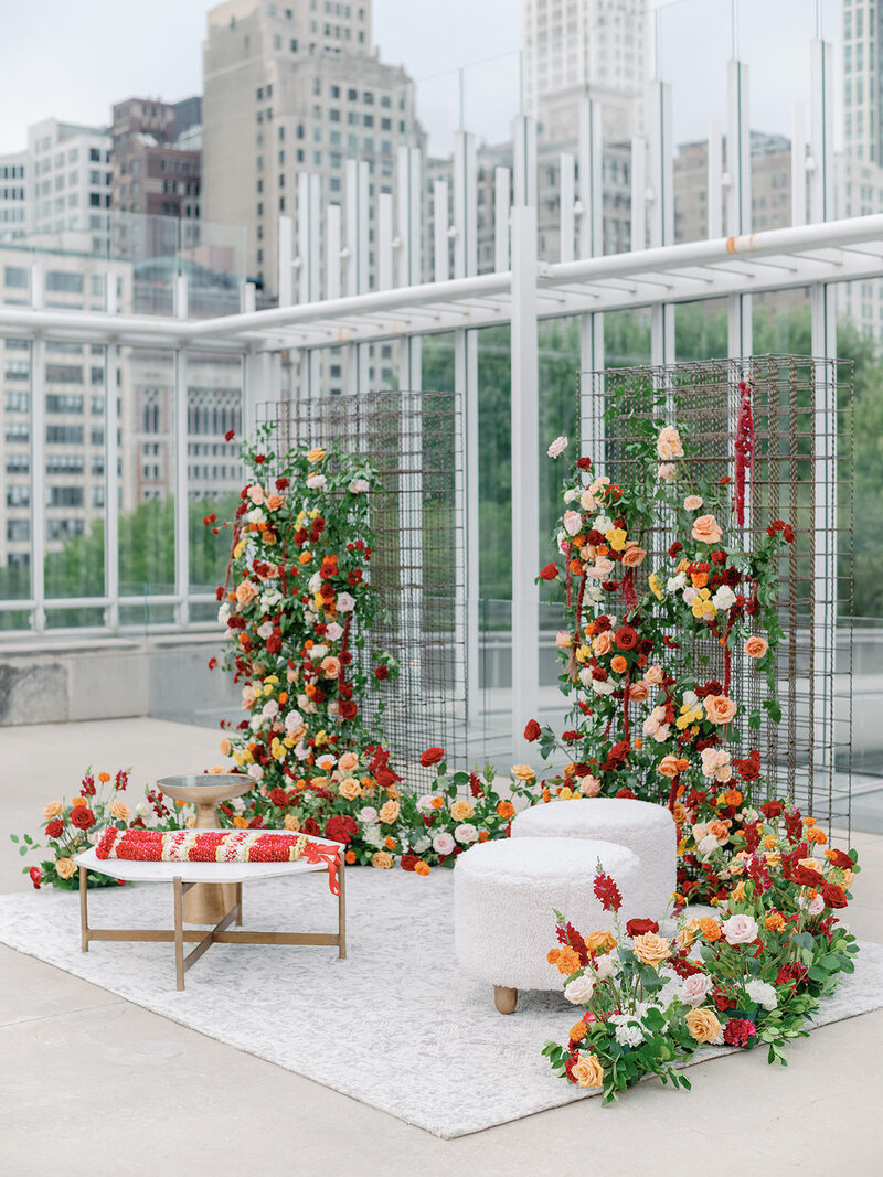 The Lounge Area of a rooftop city wedding, featuring a white table, two white ottomans, and a red , orange and peach floral backdrop