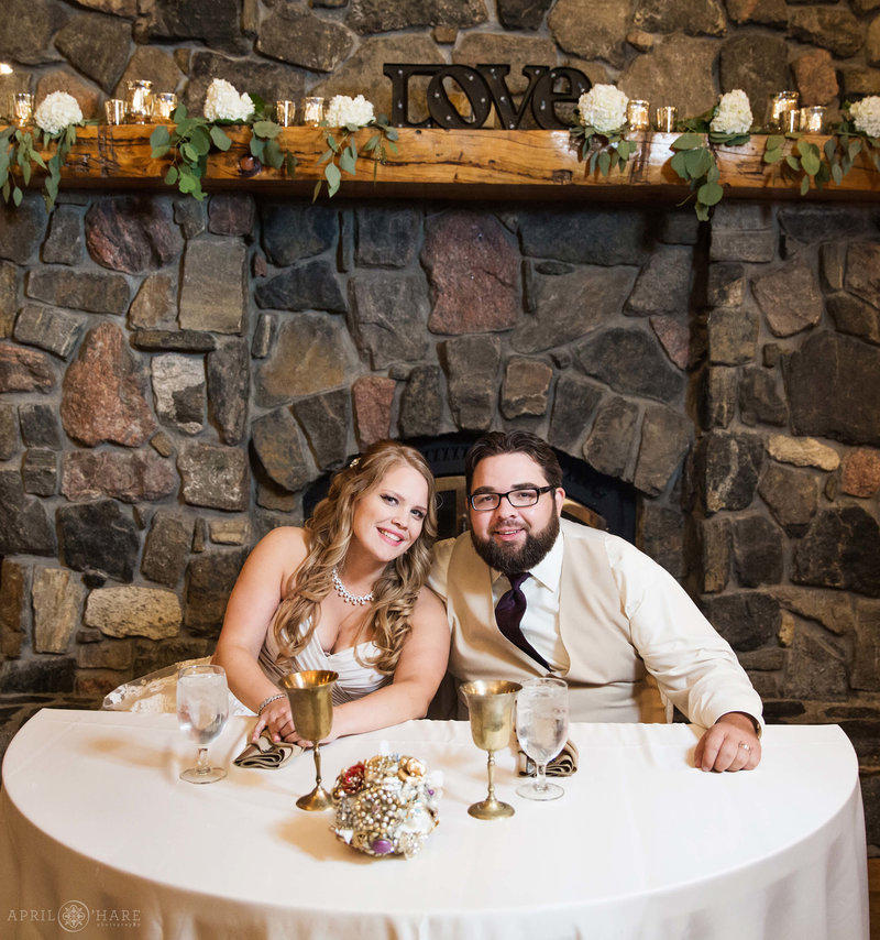 Couple seated at their sweetheart table in front of a stone fireplace inside Evergreen Lake House in Colorado