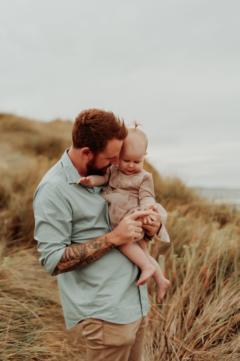 Dad stands in a field near a beach and snuggles his young daughter in her arms during a family photo session