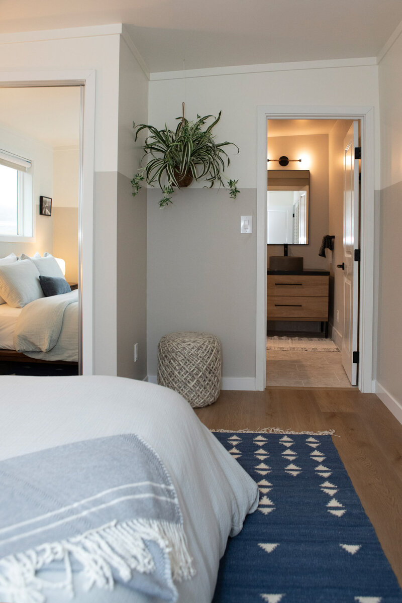 Bedroom details of a short term rental renovation showing a bed with a blue carpet beneath it, a plant hanging on the wall and a peak into the bathroom.