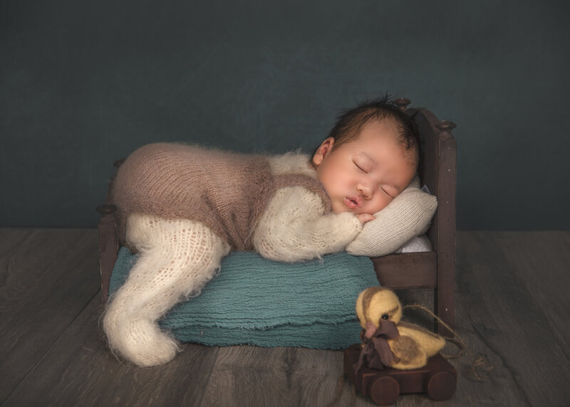 "Newborn sleeping in tiny bed with duck prop, captured in cozy portrait session"