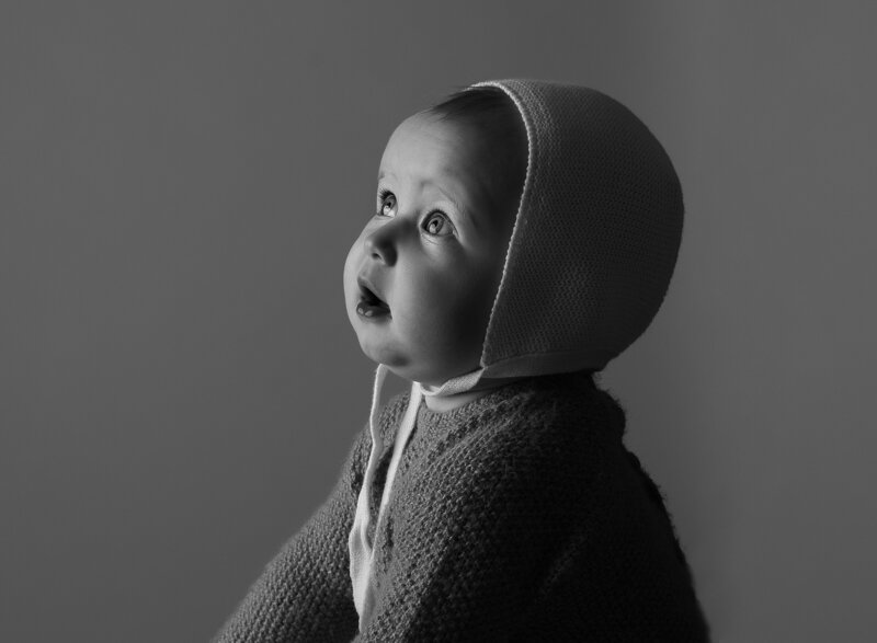 Black and white first birthday photoshoot.  Close up side profile image. Baby girl in a knit sweater and bonnet is looking upward toward the light source. Captured by best Brooklyn, NY first birthday milestone photographer Chaya Bornstein.