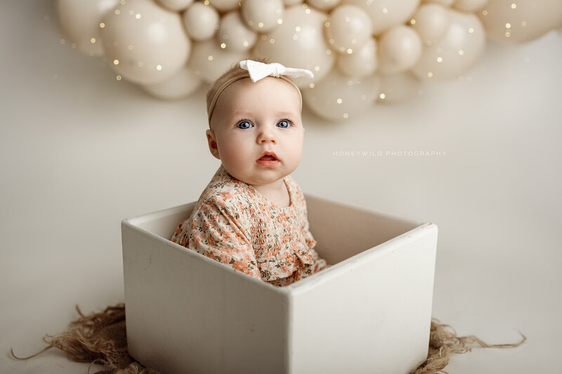 Smiling young girl in a cream dress with a bow in her hair, posing in front of a vibrant red and pink balloon backdrop during a themed portrait mini session in Grand Rapids, Michigan