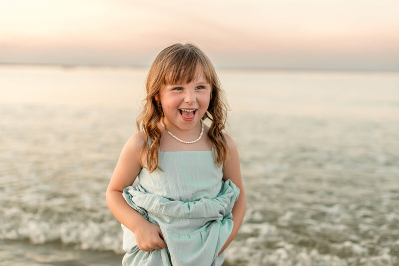 A young girl carefully lifts her dress to keep it from getting wet as she walks along the shoreline during a coastal family photo session on Tybee Island, GA.