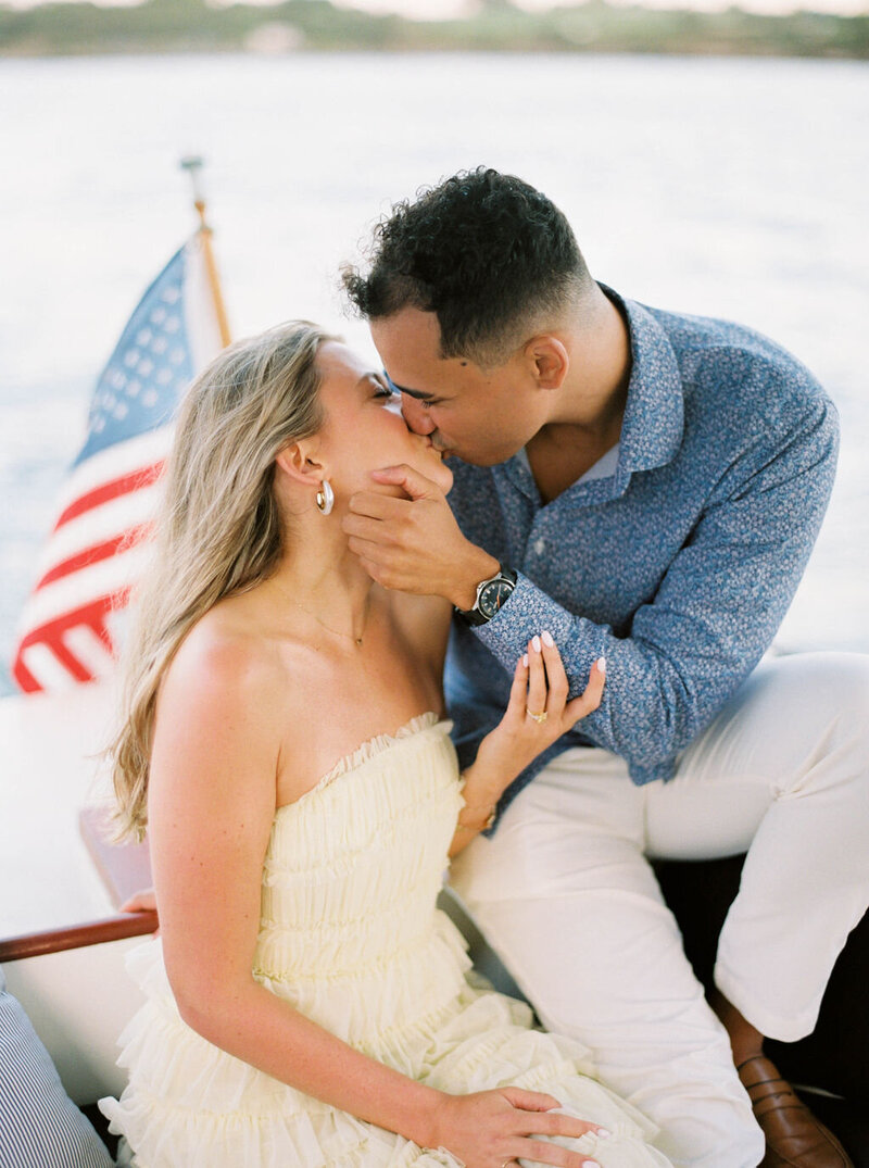 Newport Rhode Island Engagement Photo Photographer | A couple shares a romantic kiss on a boat. The woman in a strapless dress, the man in a blue shirt. An American flag waves in the background.