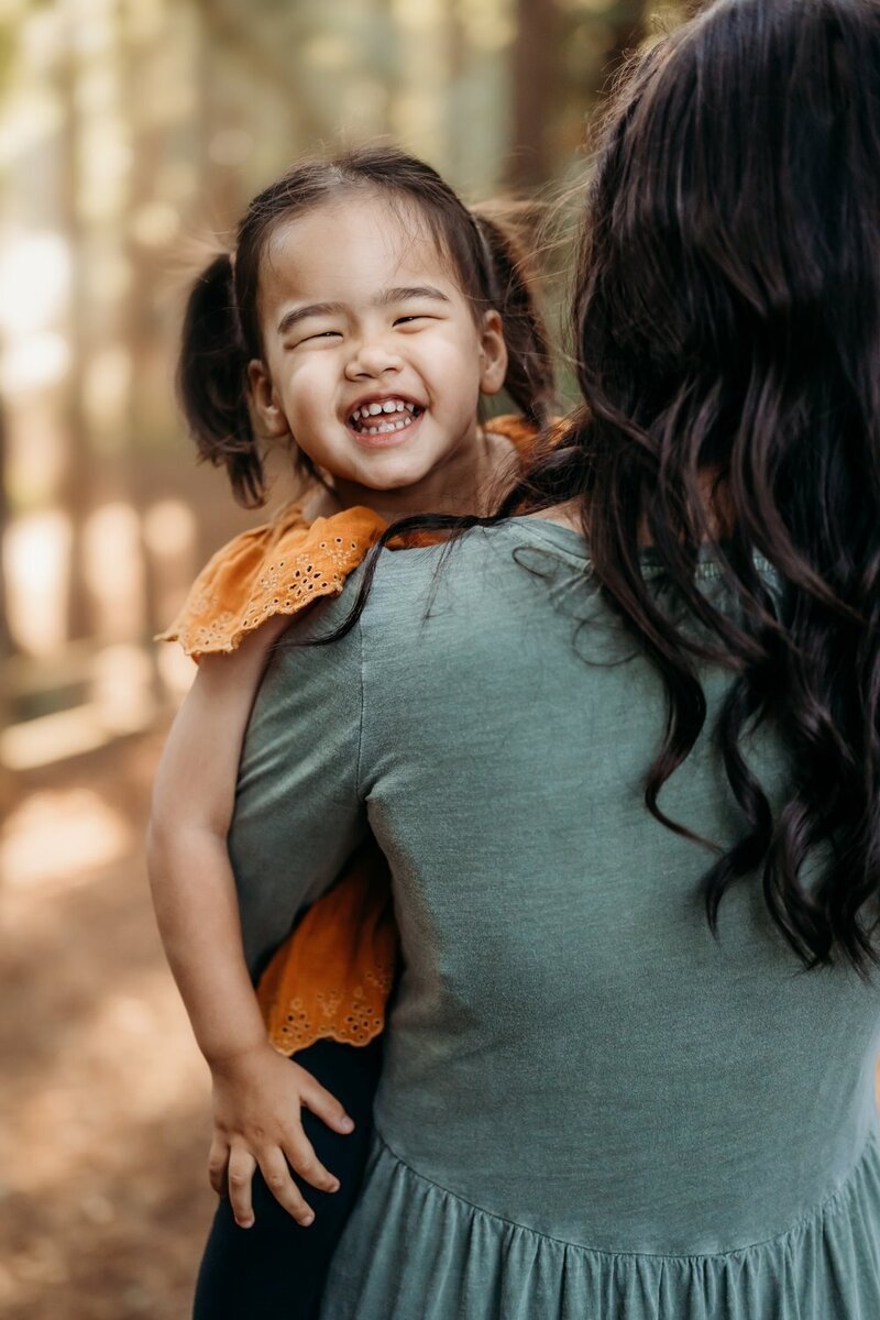 toddler smiling at the camera in her moms arms. the oakland redwoods are behind them
