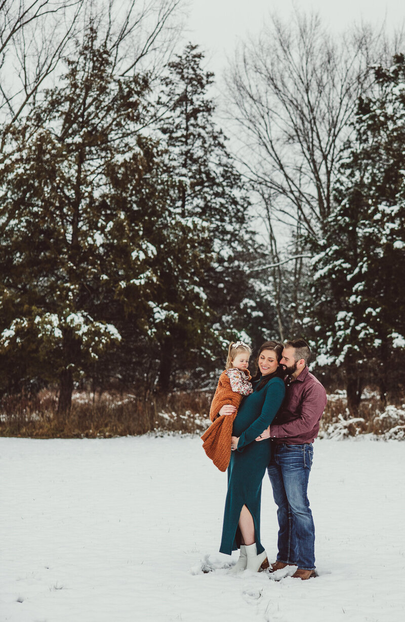 Expecting family portrait in the snow with a blanket and tall snow covered trees in the background. 