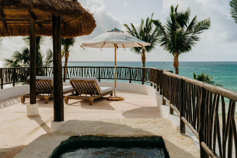 A tropical rooftop terrace overlooking the ocean, featuring two wooden lounge chairs under a large, striped umbrella. Palm trees line the edge, and a thatched pergola provides shade. The sea is visible in the background, with a clear sky above.