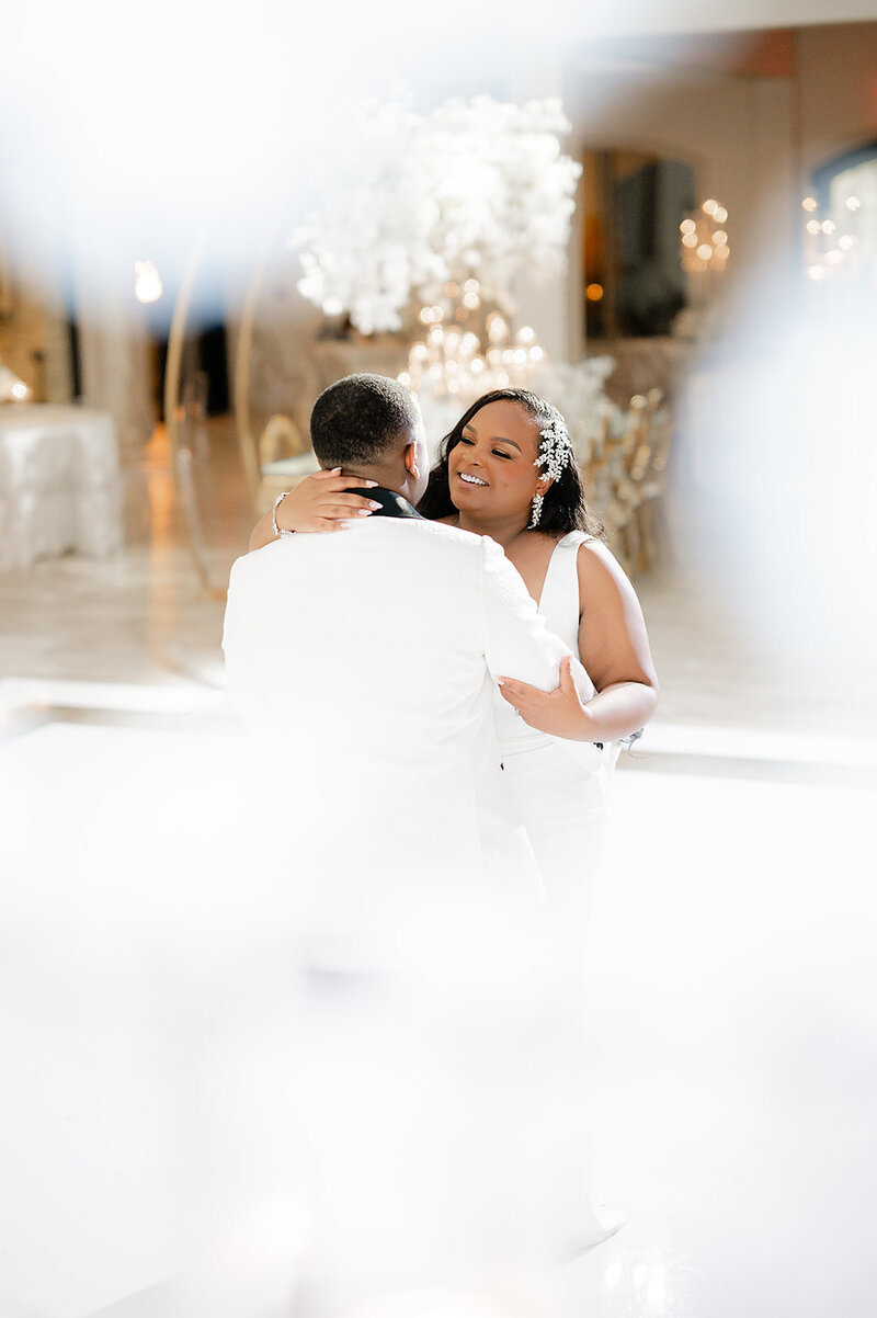 Bride smiles while the groom kisses her cheek in front of a white floral arch.