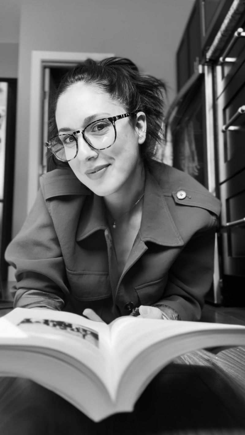 A close up shot of Hilary Rota, posed in front of her computer, with her hand under her chin, smiling and wearing glasses