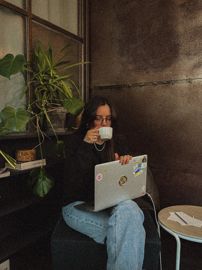 A person sits in a cozy corner, sipping coffee while working on a sticker-covered laptop. A lush plant and small table with papers add a relaxed vibe.