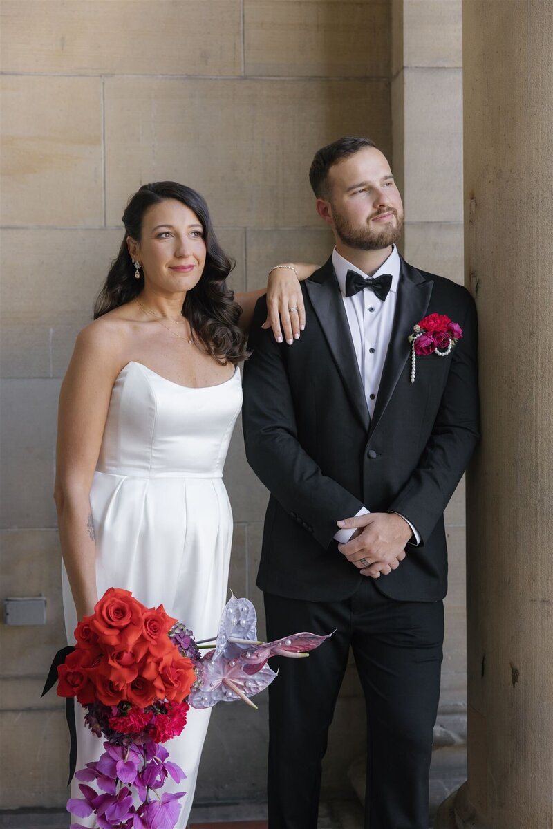 Bride and groom in front of the Line Hotel DC with the groom leaning on a pillar hands crossed and bride leaning on the shoulder of the groom relaxedly holding her bouquet at her side with the couple looking off camera