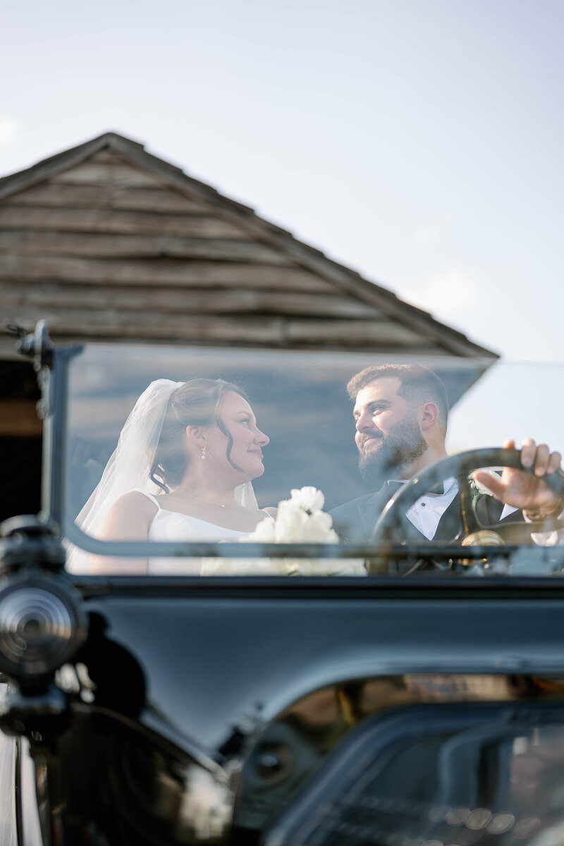 Bride and groom smiling in a vintage black car at a classic Michigan wedding.