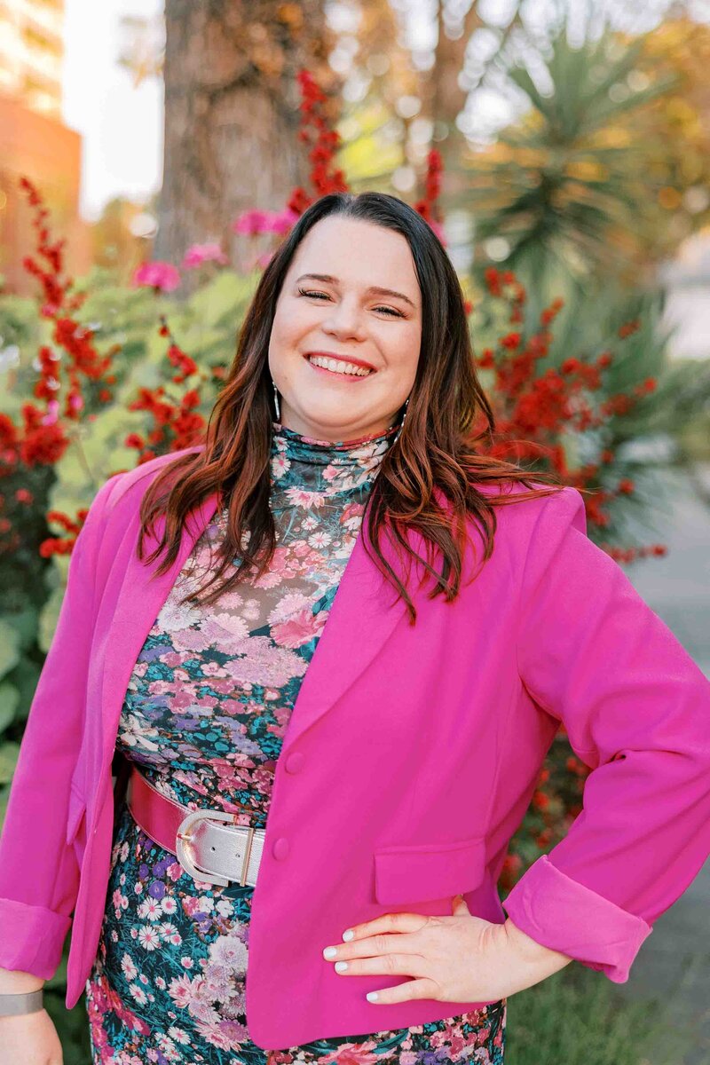 Woman wearing a hot pink blazer and floral dress and smiling at the camera. Ali Hall Art headshot