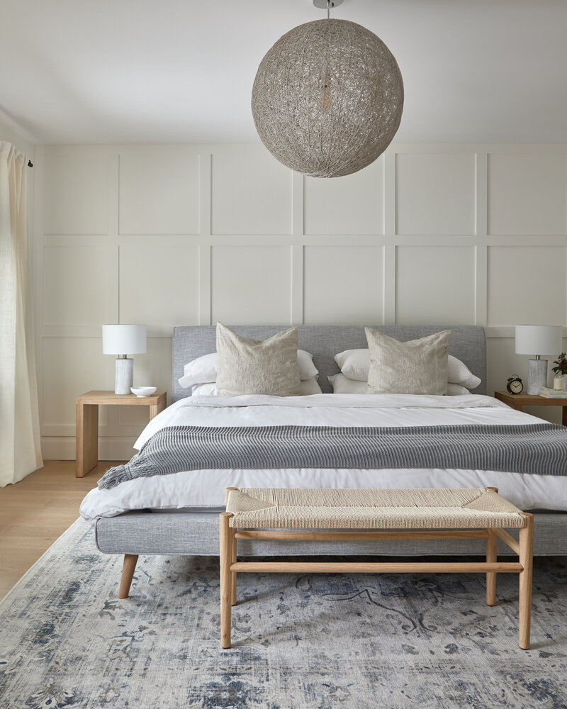 King-bed bedroom white grey fabric bed frame, light wooden floors and furniture, soft grey and off-white bedding, and a feature wall with white panelling