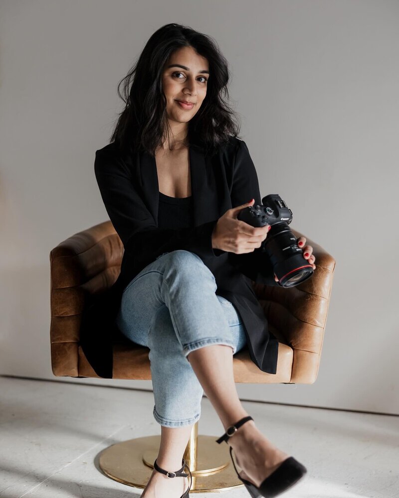 A New England lifestyle photographer, this woman in a black blazer and jeans sits on a brown chair, holding a camera. Her long dark hair cascades as she smiles slightly in the minimalistic setting with neutral tones.