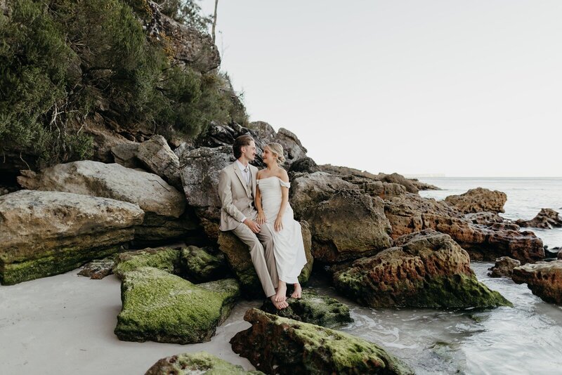 A bride and a groom sitting on the rocks at the beach