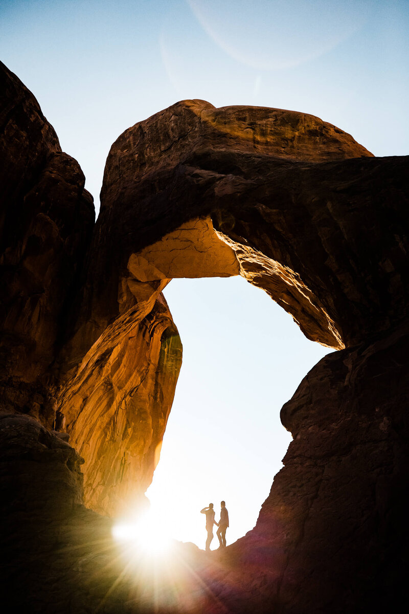 A couple stands in a red rock arch as the sun sets.