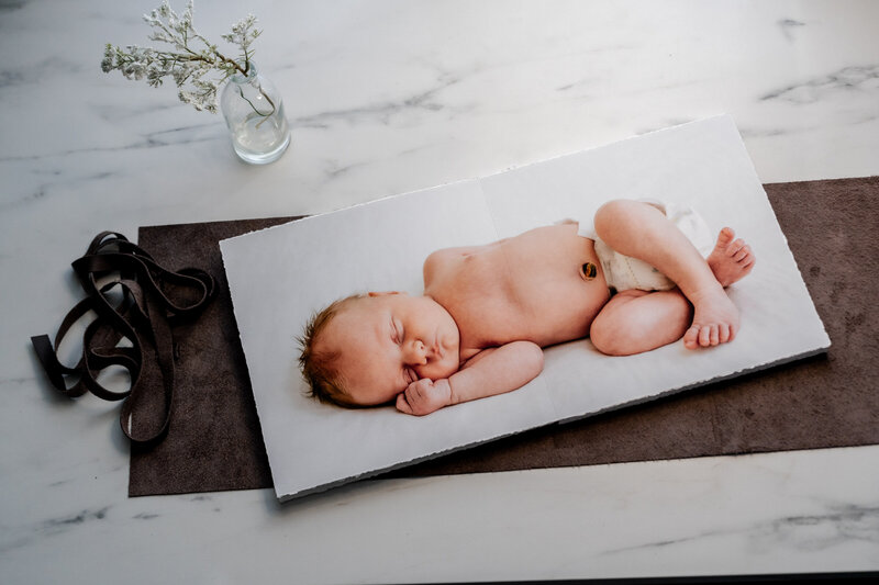 An open fine art album displaying a newborn baby sleeping peacefully on a white blanket. The album rests on a soft gray surface, with a small glass vase holding delicate white flowers beside it.