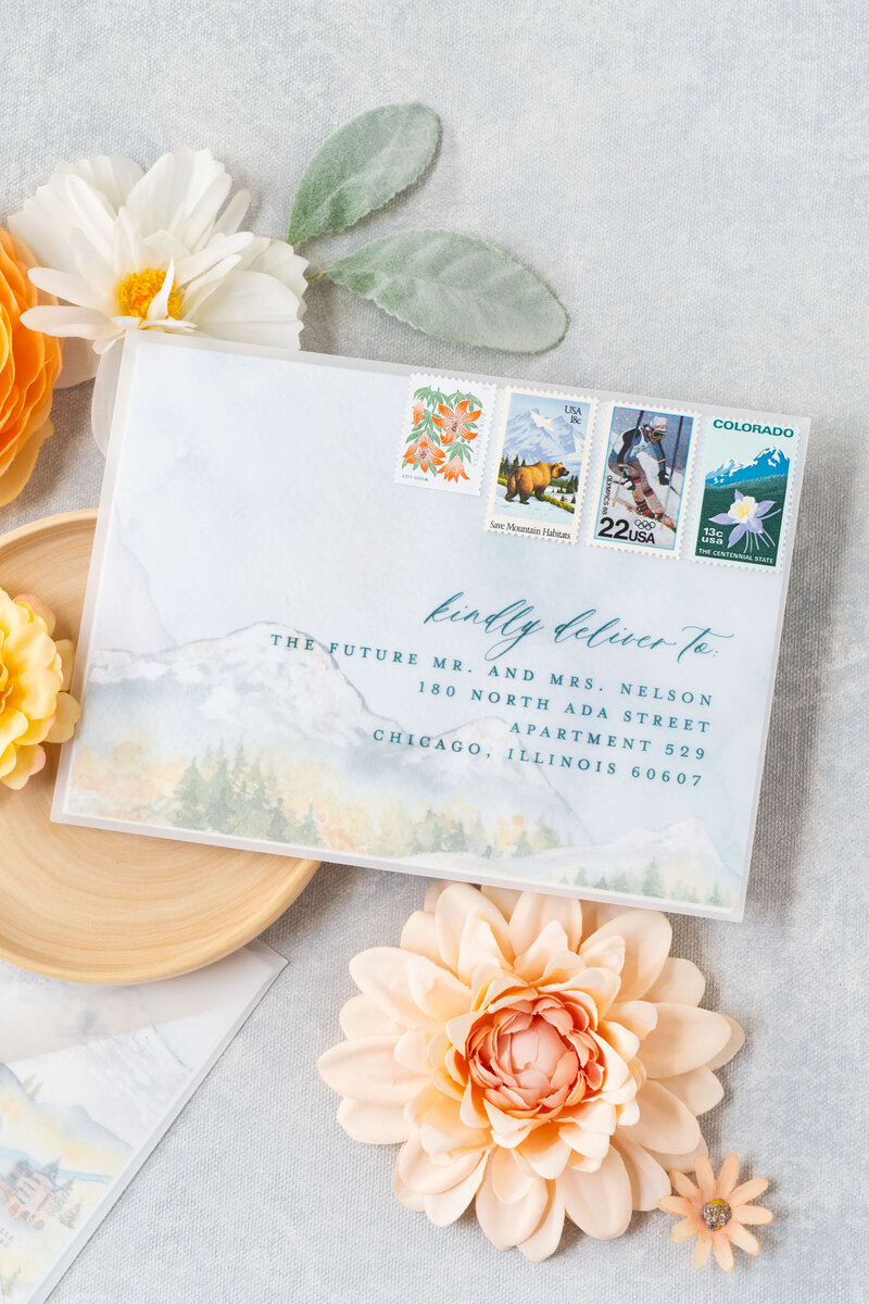 Elegant flatlay featuring a vellum envelope with a watercolor mountainscape visible through it, complemented by vintage Colorado stamps