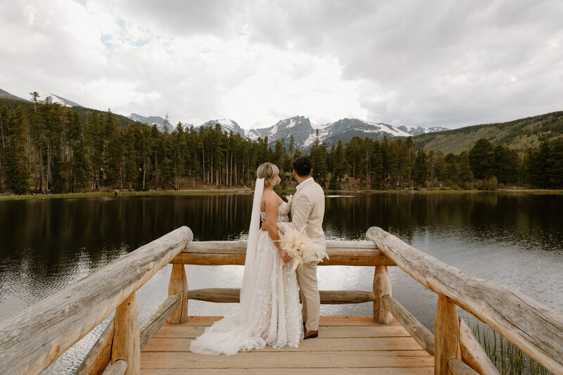 bride and groom holding hands while standing on a wooden dock overlooking a lake with mountains in the background