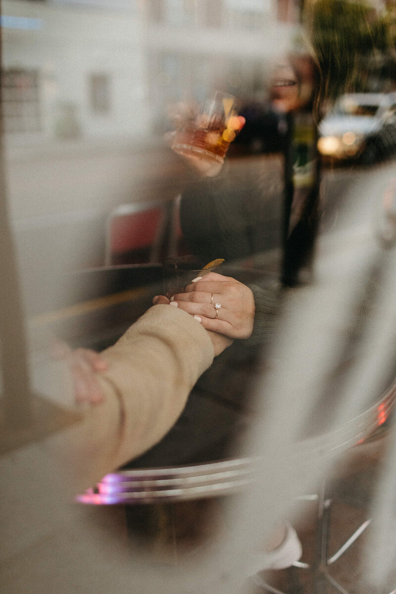Two people holding hands at a cozy cafe table, visible through the window. One person sips their drink while the other proudly displays a ring, a candid moment captured by engagement photographer, Jennifer van Son at Trinity Bellwoods.