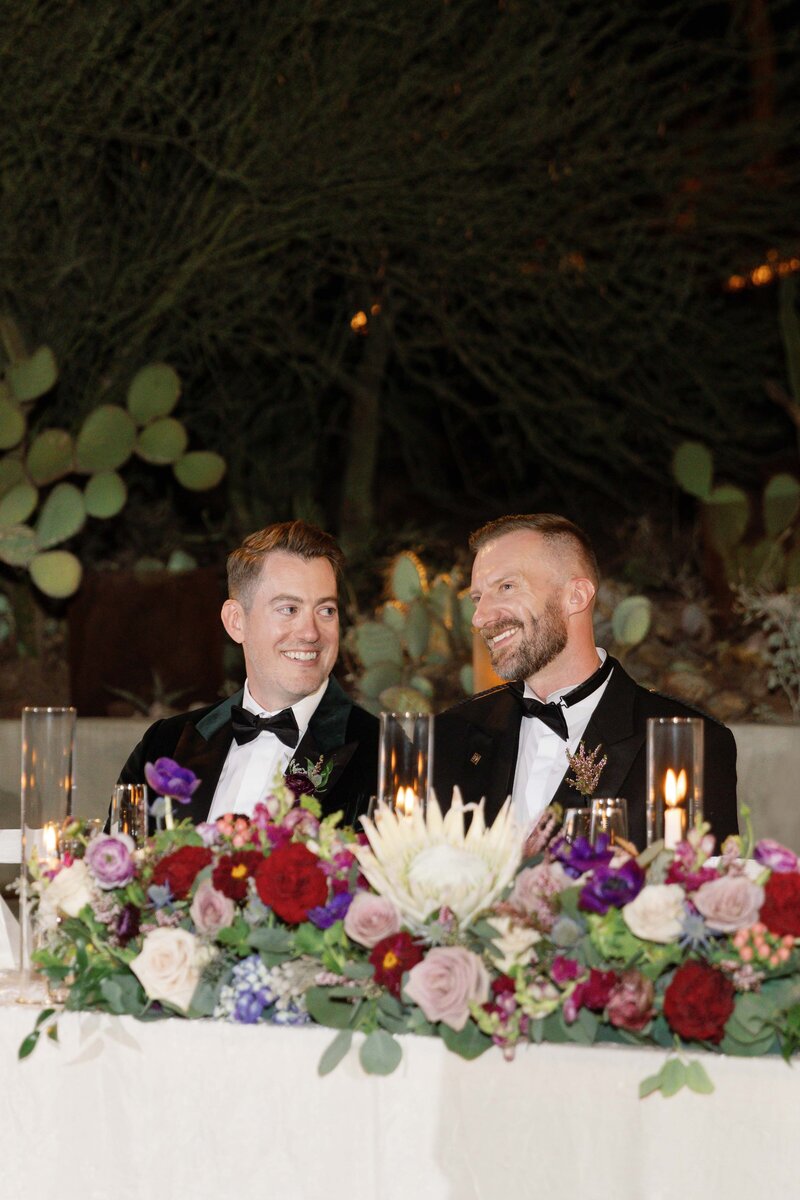 Two grooms sit at sweetheart table during their wedding and smile towards each other.