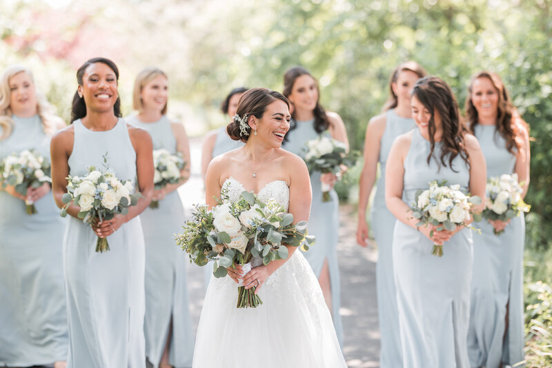 Jewish bride holding her bouquet looking back laughing while walking through the gardens at Meadowlark Botanical Gardens with her bridal party holding their bouquets smiling