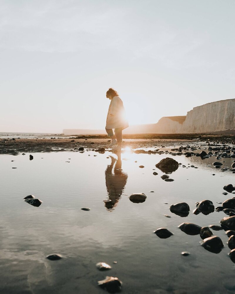 Woman walking at sunset, across the beach with rocks and mountains in the background. Peaceful.