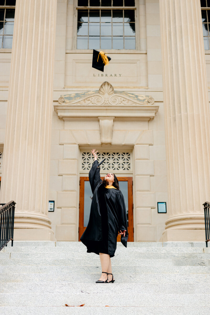 NC master’s graduate in gown and hood tossing her cap in the air on the steps of Wilson Library.