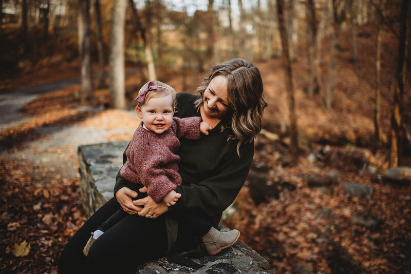 fall mommy and me session with mother sitting on a stone wall on a bridge with her toddler sitting on her lap and laughing as they are inteh woods captured by Baltimore photographers